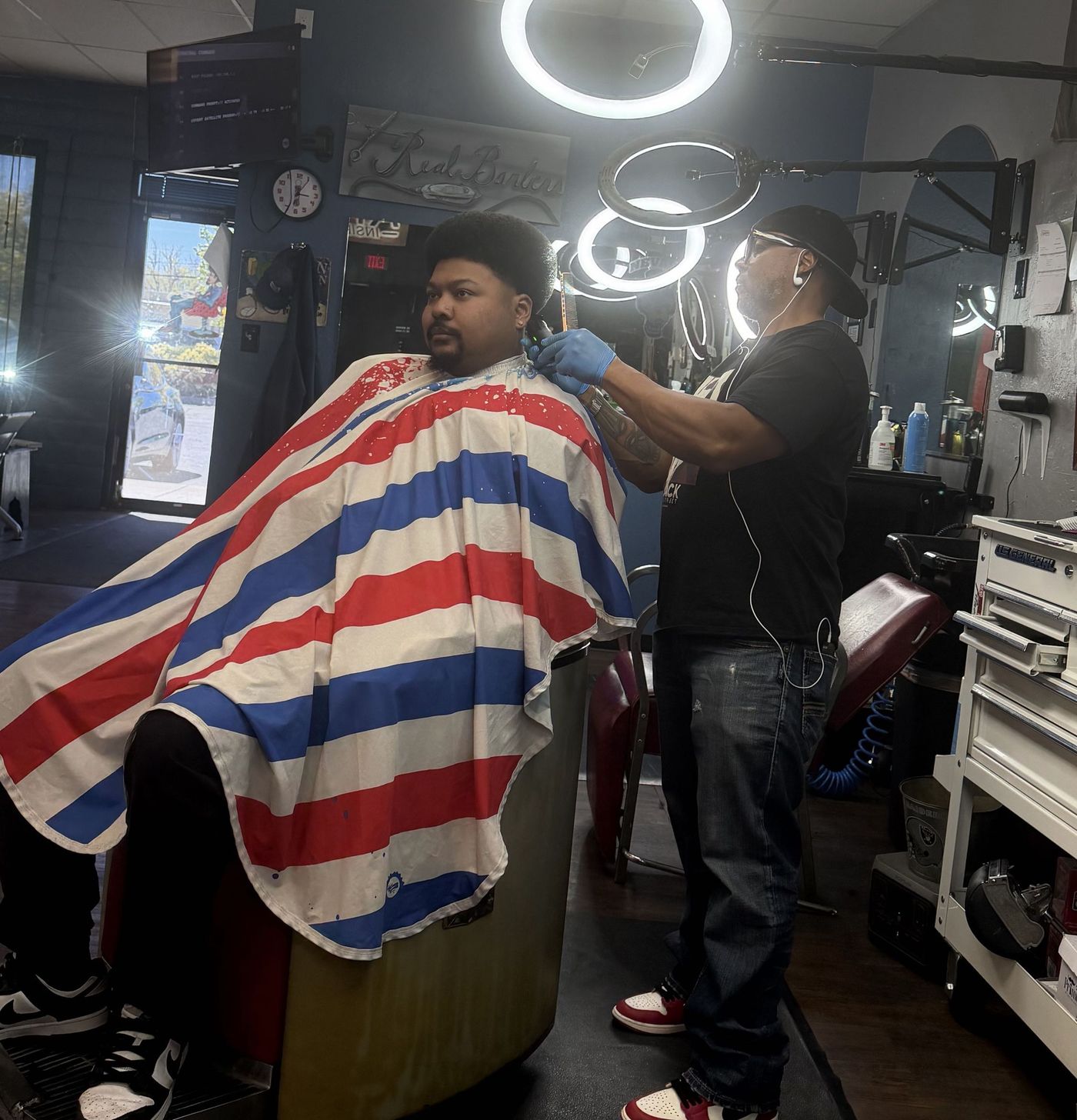 Inside Top Notch: a barber working on a client in the striped cape, vintage barber chair, ring lights overhead